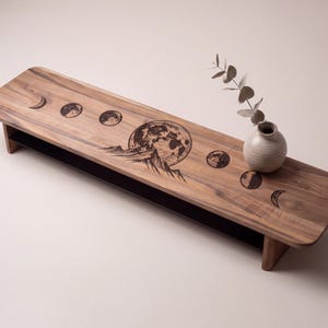 a wooden table with a decorative design depicting the phases of the moon, including the waxing and waning phases. A vase with greenery is placed on the table, adding a natural element to the scene.