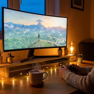 A woman is sitting at a desk, playing a video game on a large monitor. The monitor displays a scenic landscape from the video game, and the woman is holding a video game controller. The desk is decorated with various items, including a mug, a candle, and several potted plants.