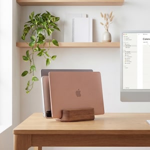 a wooden desk with a laptop, a computer monitor, and a plant. The laptop is placed on a wooden stand, and the computer monitor is on the desk. There is also a calendar on the desk, suggesting that the space is used for work and organization.