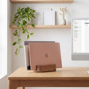a wooden desk with a laptop, a computer monitor, and a plant. The laptop is placed on a wooden stand, and the computer monitor is on the desk. There is also a calendar on the desk, suggesting that the space is used for work and organization.