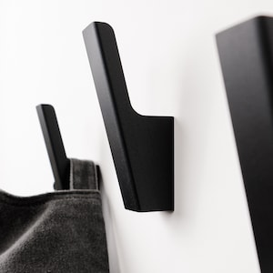Pode incluir: Close-up de três ganchos de parede pretos montados numa parede branca. Os ganchos têm um design moderno e minimalista, com uma base retangular e um gancho curvo. Um item de tecido cinzento está pendurado num gancho.