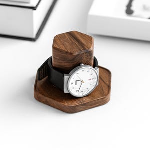 Pode incluir: Um relógio de pulso moderno com mostrador branco e pulseira de couro preta repousa sobre um suporte de relógio de madeira. O suporte é feito de madeira marrom escura e tem um design geométrico. O relógio tem ponteiros prateados e uma caixa prateada.