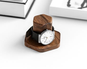 Op de afbeelding: Een modern polshorloge met een witte wijzerplaat en een zwarte leren band rust op een houten horlogestandaard. De standaard is gemaakt van donkerbruin hout en heeft een geometrisch ontwerp. Het horloge heeft zilverkleurige wijzers en een zilverkleurige kast.