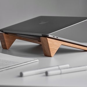 May include: A laptop computer sits on a wooden stand. The stand is made of light-coloured wood and has a minimalist design. Two white pens and a sheet of paper are in the foreground.
