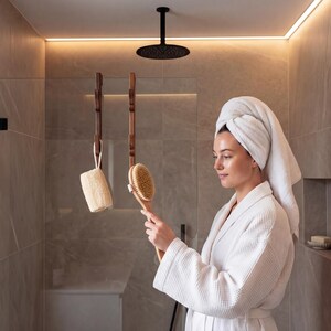 A woman in a white bathrobe stands in a modern bathroom, holding a wooden brush and looking at a mirror.