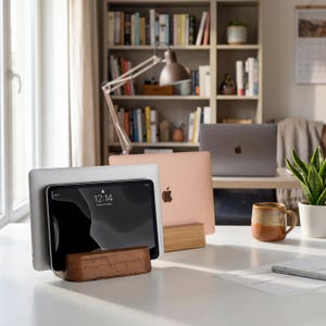 Puede incluir: Dos soportes de madera sostienen dispositivos electrónicos en un escritorio blanco. Un soporte de nogal oscuro sostiene una tableta plateada que muestra las 12:14, mientras que un soporte de roble más claro sostiene un portátil.