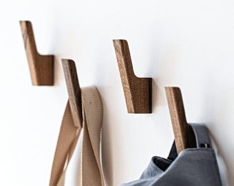May include: Four individual wall-mounted hooks crafted from dark brown wood with a smooth, angled design. Each hook features a vertical post with a slight upward tilt, providing a functional surface for hanging items like fabric straps and bags.