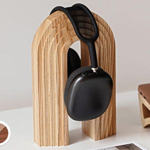 May include: A wooden headphone stand in a light brown finish, holding a pair of black headphones. The stand has an arched design with a layered texture. The headphones are black with a mesh headband and earcups. The stand is on a white surface.