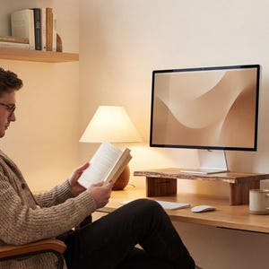 A man is sitting at a desk, reading a book and looking at a computer monitor.