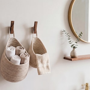 a bathroom setting with two woven baskets hanging on the wall, each containing rolled towels. A round mirror is mounted above the baskets, and a wooden shelf is visible below the mirror. A small potted plant is placed on the shelf, adding a touch of greenery to the space.