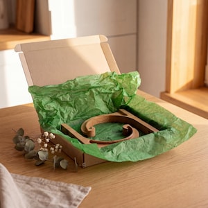 A wooden object, possibly a piece of furniture, is displayed in a cardboard box with green tissue paper, placed on a wooden table.