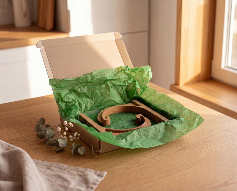 A wooden object, possibly a piece of furniture, is displayed in a cardboard box with green tissue paper, placed on a wooden table.