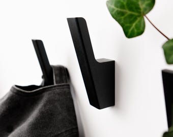 May include: A set of matte black wall-mounted hooks with an angled, geometric silhouette. The hooks are attached to a white surface, with one hook supporting a dark gray fabric item and another positioned near a green leafy plant.