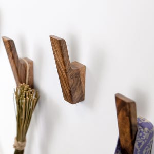 May include: Three wall-mounted coat hooks crafted from dark brown wood with a visible natural grain. Each hook features an angled, upward-pointing design. One hook holds a bundle of dried grass, and another holds a folded purple patterned fabric.