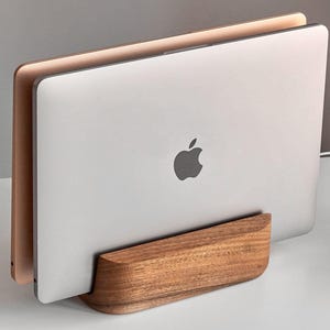 May include: A silver laptop with the Apple logo is held in a wooden stand. The stand is made of a light brown wood and has a curved design. A second laptop with a gold finish is also visible.