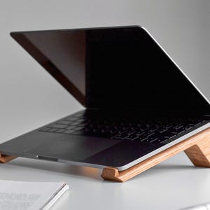 May include: A laptop computer is elevated on a wooden stand. The stand is made of light brown wood and has a triangular shape. The laptop is closed and has a black screen.