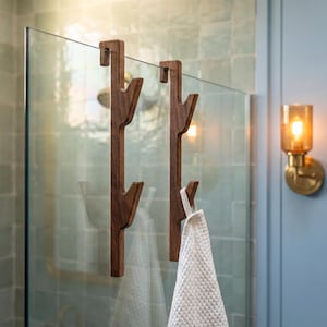a bathroom with a glass shower door, a wooden towel rack with two towel hooks, and a light fixture mounted on the wall.