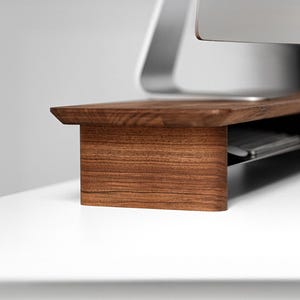 Puede incluir: Un soporte de monitor de madera marrón oscuro con una superficie superior horizontal lisa y dos soportes laterales verticales. El soporte presenta un patrón de veta de madera visible y está diseñado para elevar una pantalla de ordenador.