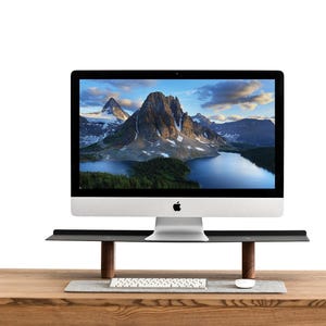 Könnte beinhalten: Ein silberner iMac-Computer mit einer Berglandschaft auf dem Bildschirm steht auf einem schwarzen Monitorständer mit Holzbeinen. Eine weiße Tastatur und Maus befinden sich auf dem Holzschreibtisch.