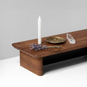 May include: A dark brown wooden shelf displaying a white candle, a brass dish, a clear crystal, and lavender. The shelf has a black underside. The background is white and grey. The shelf is approximately 76 cm long.