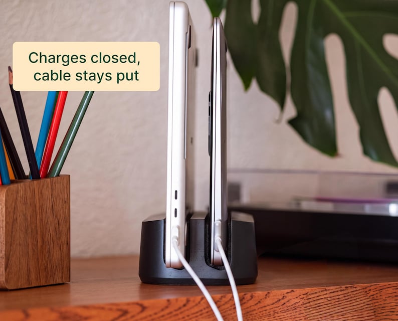 two laptops connected to a charging station, with a wooden desk and a plant in the background.