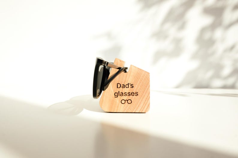 A pair of black sunglasses is placed on a wooden stand with the text "Dad's glasses on" engraved on it.
