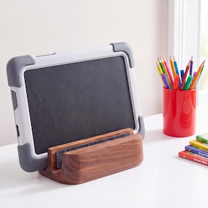 May include: A tablet with a gray protective case rests in a horizontal wooden stand. Beside it, a red cylindrical pencil holder contains various colored pencils, and a small stack of school workbooks lies on the white surface.