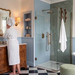 A woman in a white bathrobe stands at a bathroom vanity, looking at herself in the mirror. The bathroom has a blue and white color scheme, with a glass-enclosed shower, a sink, and a bathtub.
