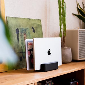 May include: A silver laptop with a black Apple logo is displayed in a black stand on a wooden surface. The laptop is open, revealing stickers on the inside. A green painting and a speaker are in the background.