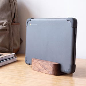 Op de afbeelding: Een donkergrijze Chromebook-laptop met afgeronde hoekbeschermers wordt rechtop gehouden door een rechthoekige standaard van massief hout met een zichtbare natuurlijke houtnerf. Het veelkleurige Chromebook-logo staat linksboven op het deksel.
