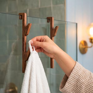 A hand holding a white towel in front of a glass shower door with wooden hooks.