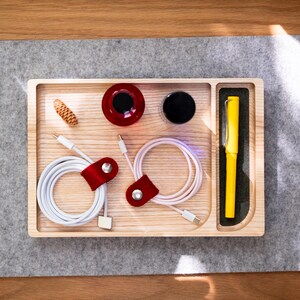May include: A wooden desk organizer with compartments holding a yellow pen, white charging cables with red leather cable organizers, a red and black object, a black object, and a small pinecone. The organizer sits on a gray felt surface.