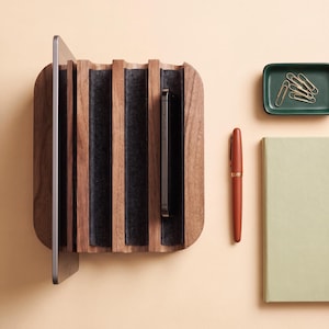 Op de afbeelding: Een houten bureau-organizer met verticale sleuven bekleed met donkergrijs vilt, waarin een tablet en een smartphone staan. Daarnaast liggen een donkergroen bakje met gouden paperclips, een terracotta vulpen en een lichtgroen notitieboek.