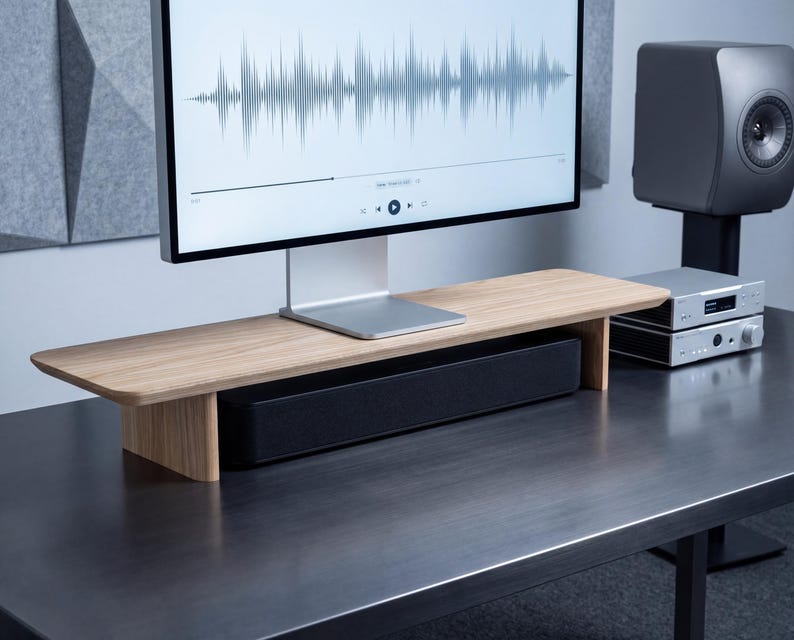 a modern, minimalist workspace with a large flat-screen monitor displaying a sound waveform, a wooden desk with a black speaker system, and a gray speaker system.