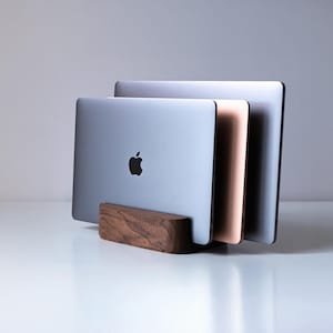 May include: A dark brown wooden vertical laptop stand holding three closed laptops of varying colors, including silver, rose gold, and space gray. The stand features a smooth, rounded rectangular base with a visible wood grain texture.