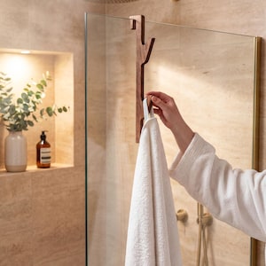 A person's hand is holding a white towel in front of a glass shower door, with a wooden towel rack attached to the glass.
