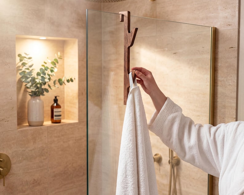 A person's hand is holding a white towel in front of a glass shower door, with a wooden towel rack attached to the glass.