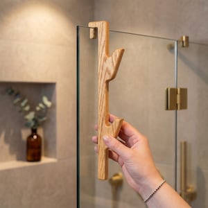 A hand is holding a wooden handle with a unique design, positioned in front of a glass shower door.