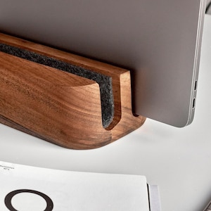 May include: A dark brown wooden laptop stand with a smooth, curved design and a dark gray felt lining in the center slot. The stand holds a silver laptop in an upright position, showcasing the natural wood grain and the contrast of the felt material.