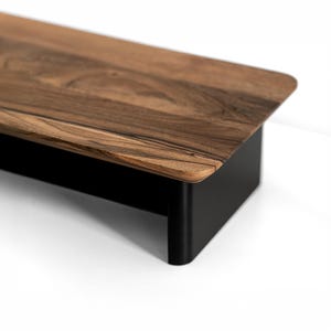 May include: A rectangular monitor stand featuring a smooth, dark brown wood grain top surface with visible natural patterns. The stand is supported by a solid, matte black metal base with rounded corners, providing a stable elevated platform.