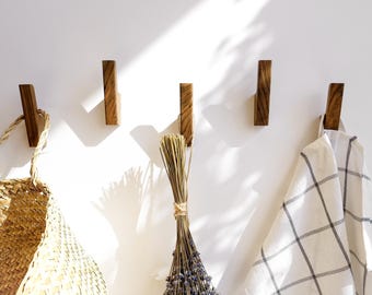 May include: Five rectangular wooden wall hooks mounted in a horizontal row. A woven straw basket hangs from the first hook, a bundle of dried lavender hangs from the third hook, and a white cloth with a grey grid pattern hangs from the fifth hook.