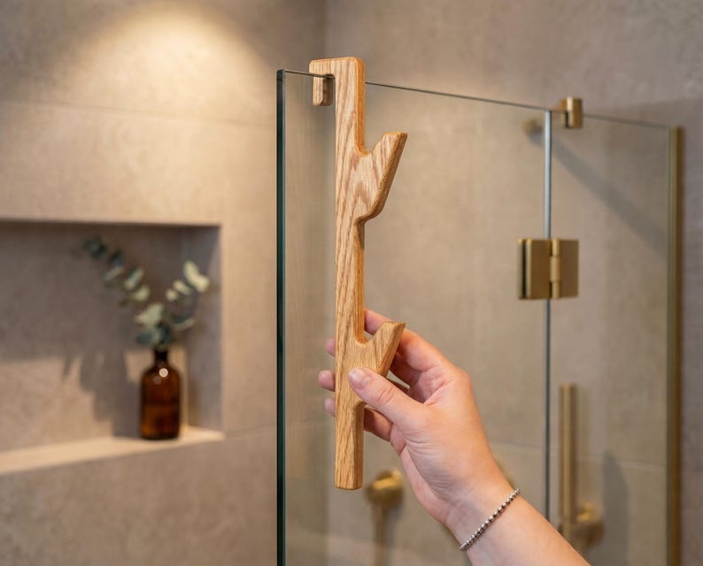 A hand is holding a wooden handle with a unique design, positioned in front of a glass shower door.
