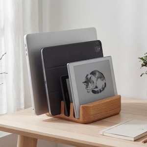 May include: A wooden desk organiser holds a silver laptop, a black tablet, and a digital reader with a cat illustration. The organiser is made of light-coloured wood and sits on a wooden desk. A small bonsai tree is to the right.