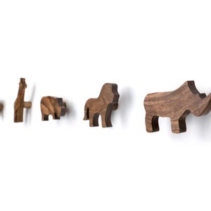 Op de afbeelding: Houten wandhaken in de vorm van dieren. De set bevat een vierkante haak, een giraffe, een nijlpaard, een leeuw en een neushoorn. Elke haak is uit hout gesneden en heeft een natuurlijke houtnerf. De haken zijn op een witte muur gemonteerd.