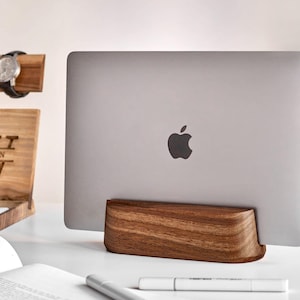 May include: A silver laptop with an Apple logo is held upright by a solid dark brown wooden stand. The stand has a smooth, curved design and a visible wood grain pattern. The laptop is positioned vertically, showcasing its metallic finish.
