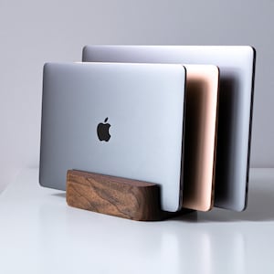 May include: A wooden laptop stand holding three laptops, silver and rose gold in colour. The stand is made of dark wood and has a rectangular shape. The Apple logo is visible on the front of the silver laptop.