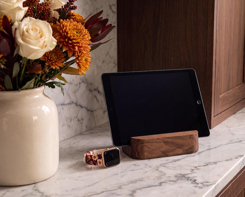 a tablet resting on a wooden stand, with a vase of white and orange flowers placed next to it on a marble countertop.