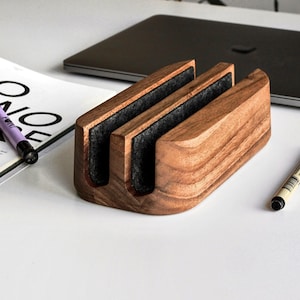 May include: A wooden desk organiser with two slots lined with dark grey felt. The organiser is made of light brown wood with visible grain. A purple pen and a black pen are next to the organiser. A laptop is in the background.