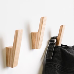 Puede incluir: Tres ganchos de pared de madera de color claro con un diseño cónico y angulado. Los ganchos están montados sobre una superficie lisa, y un objeto de tela negra con una hebilla de metal plateado cuelga del gancho derecho, mostrando la veta.