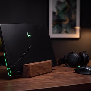 a sleek black laptop with a green alien logo on the back, placed on a wooden desk. The laptop is accompanied by a mouse and headphones, suggesting a workspace setup for gaming or multimedia purposes.